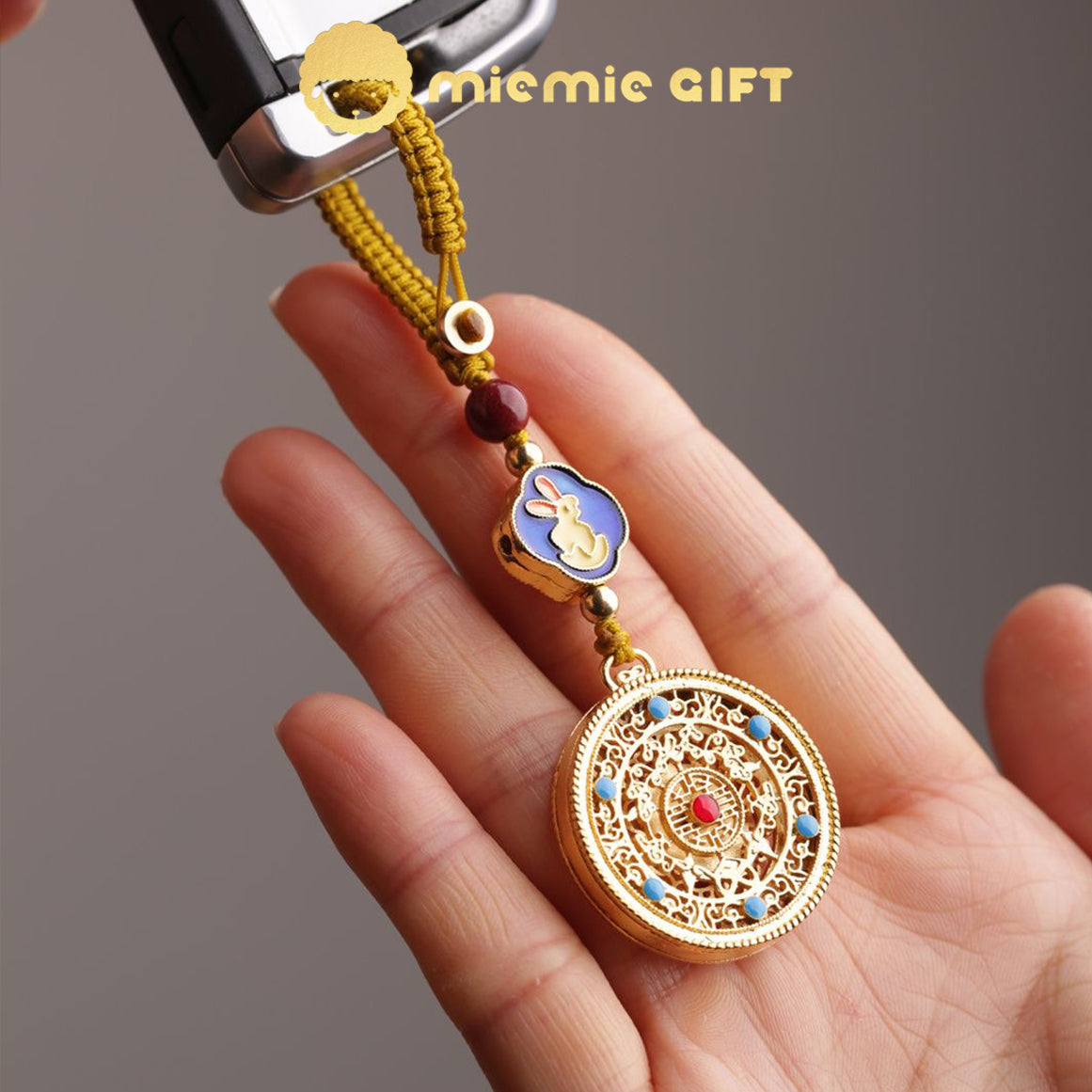 A 12 Zodiac Lucky Charm Pendant attached to a car key, held in hand. Features a golden alloy spinning disk, a vibrant rabbit zodiac symbol, hand-braided cord, and faux cinnabar bead. A stylish and meaningful accessory for luck and prosperity.