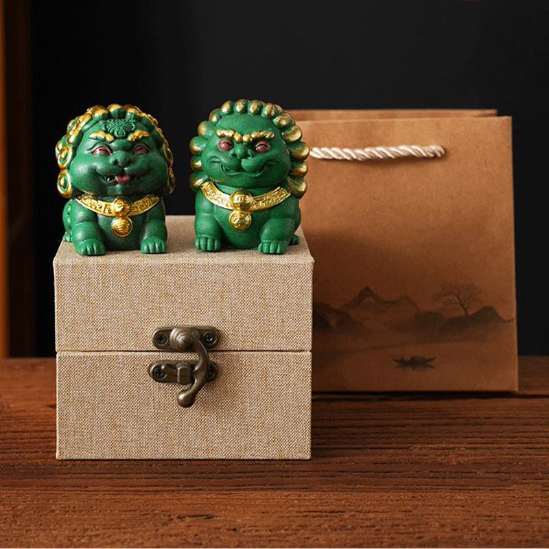 A pair of green mini stone lions, the Fortune Lion and Peace Lion, adorned with golden accents, displayed on a beige gift box with a decorative paper bag in the background, symbolizing protection and prosperity.