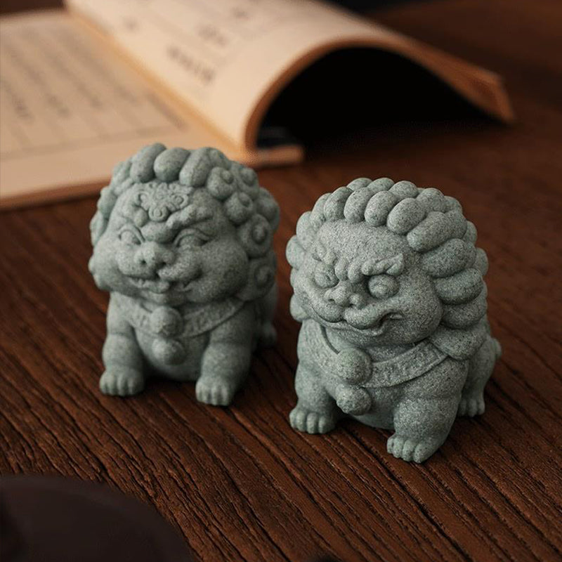 A pair of green sandstone mini stone lion ornaments, the Fortune Lion and Peace Lion, displayed on a wooden surface with an open book in the background, symbolizing protection and prosperity.