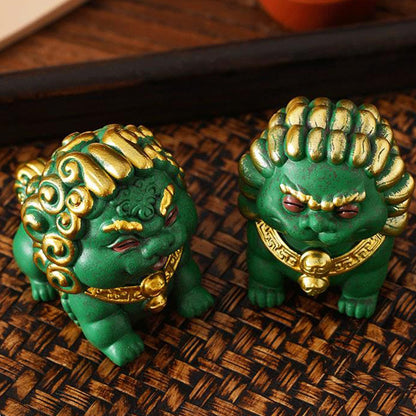 Two mini stone lion ornaments in vibrant green with intricate gold accents, displayed on a woven surface. Their expressive faces and ornate details reflect traditional Chinese craftsmanship, symbolizing wealth, protection, and harmony.