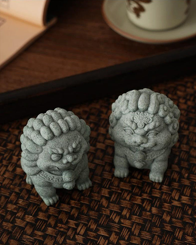 A pair of green sandstone mini stone lions, the Peace Lion and Fortune Lion, displayed on a woven surface with a teacup and book in the background, symbolizing traditional charm and protection.