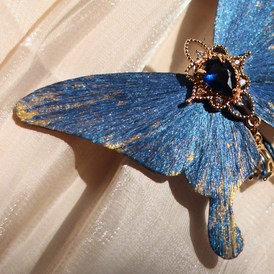 Close-up of a handcrafted deep ocean blue velvet butterfly brooch by Chujiu Mountain Atelier, featuring golden accents, a vibrant blue crystal centerpiece, and shimmering textures inspired by the ocean's elegance.