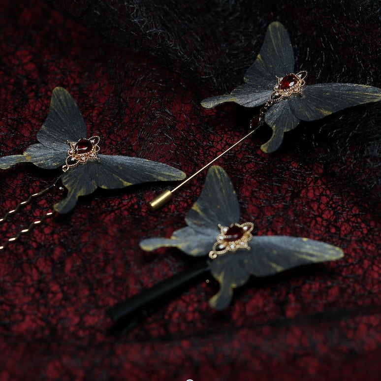 A set of three gray butterfly velvet flower hairpins adorned with red gemstones and golden accents, displayed on a textured black lace background with deep red undertones.






