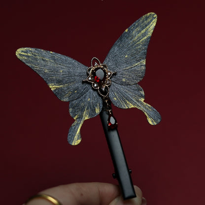 Hand holding a gray butterfly velvet flower hair clip featuring an elegant red gemstone centerpiece and dangling embellishments, set against a rich red background.