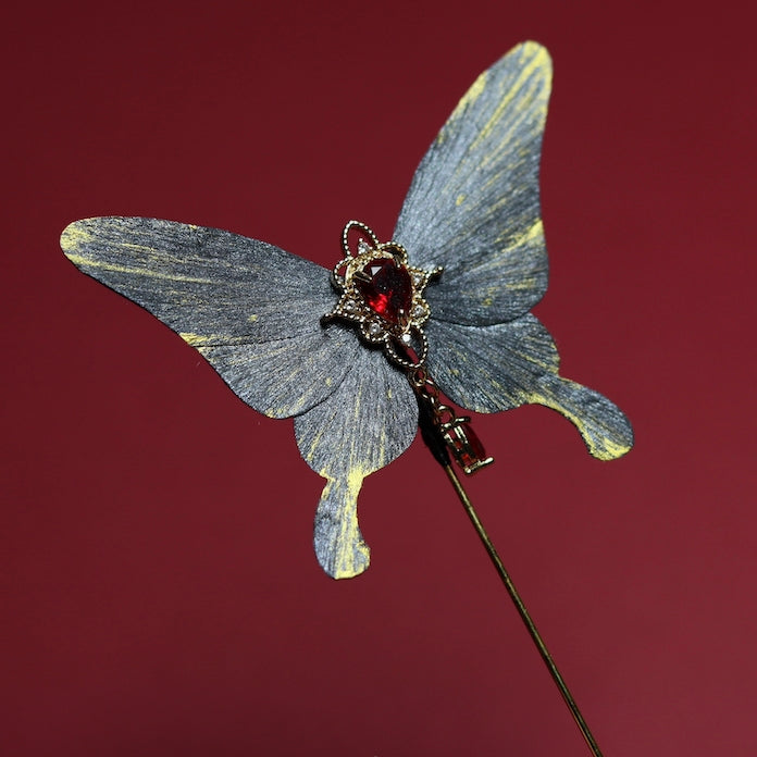 A close-up of a single gray butterfly velvet flower hairpin featuring a vivid red crystal centerpiece and intricate gold accents, set against a rich red background.