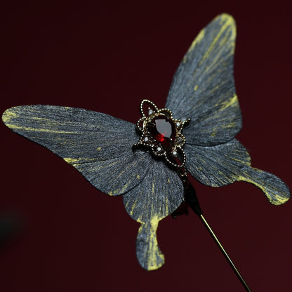 Close-up of a gray butterfly velvet flower hairpin adorned with a striking red crystal centerpiece and intricate gold detailing, set against a deep red background.