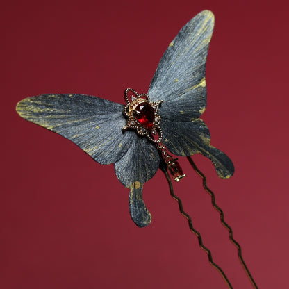 A stunning gray butterfly-shaped velvet hairpin adorned with a vibrant red gemstone and delicate gold highlights, displayed on a crimson backdrop