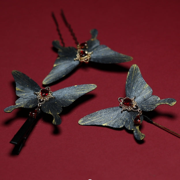 A set of three gray butterfly velvet flower hairpins, each featuring a radiant red crystal centerpiece and delicate gold accents, arranged on a deep red background.