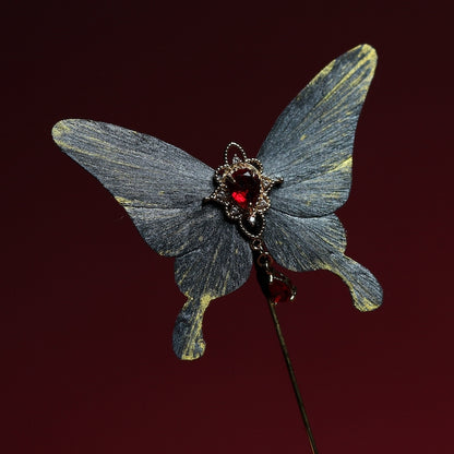 A delicate gray butterfly velvet flower hairpin adorned with a sparkling red gemstone centerpiece and dangling accents, displayed against a deep red background.