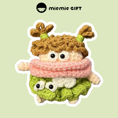 Handcrafted knit earphone case featuring a cute character with curly brown hair tied in pigtails, a pink scarf, and a green skirt. The playful design includes large eyes and unique details, offering a fun and quirky way to protect your AirPods.