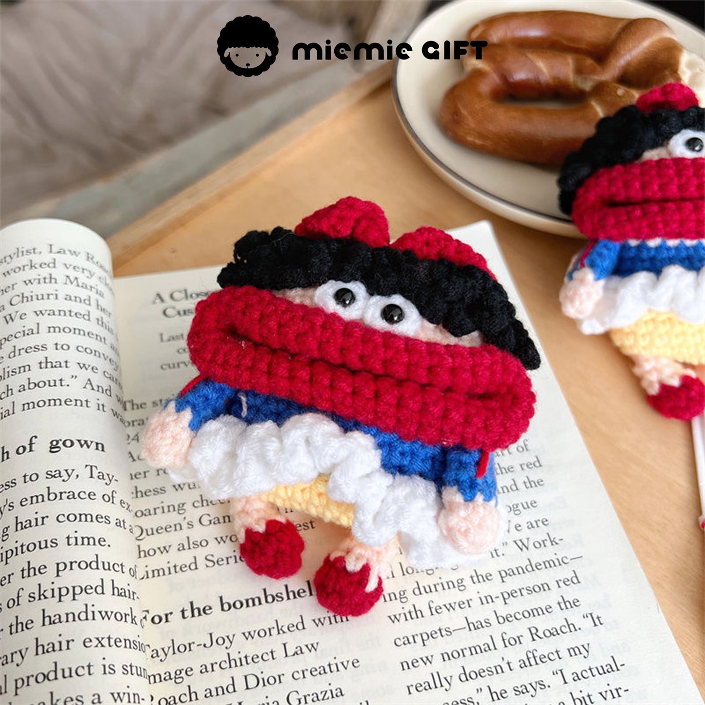 Handcrafted Snow White knit earphone case placed next to a book and a bagel. The case features a playful Snow White character with red, blue, and white colors, complete with black curly hair and a red bow, designed to protect your AirPods with a fun, whimsical look.