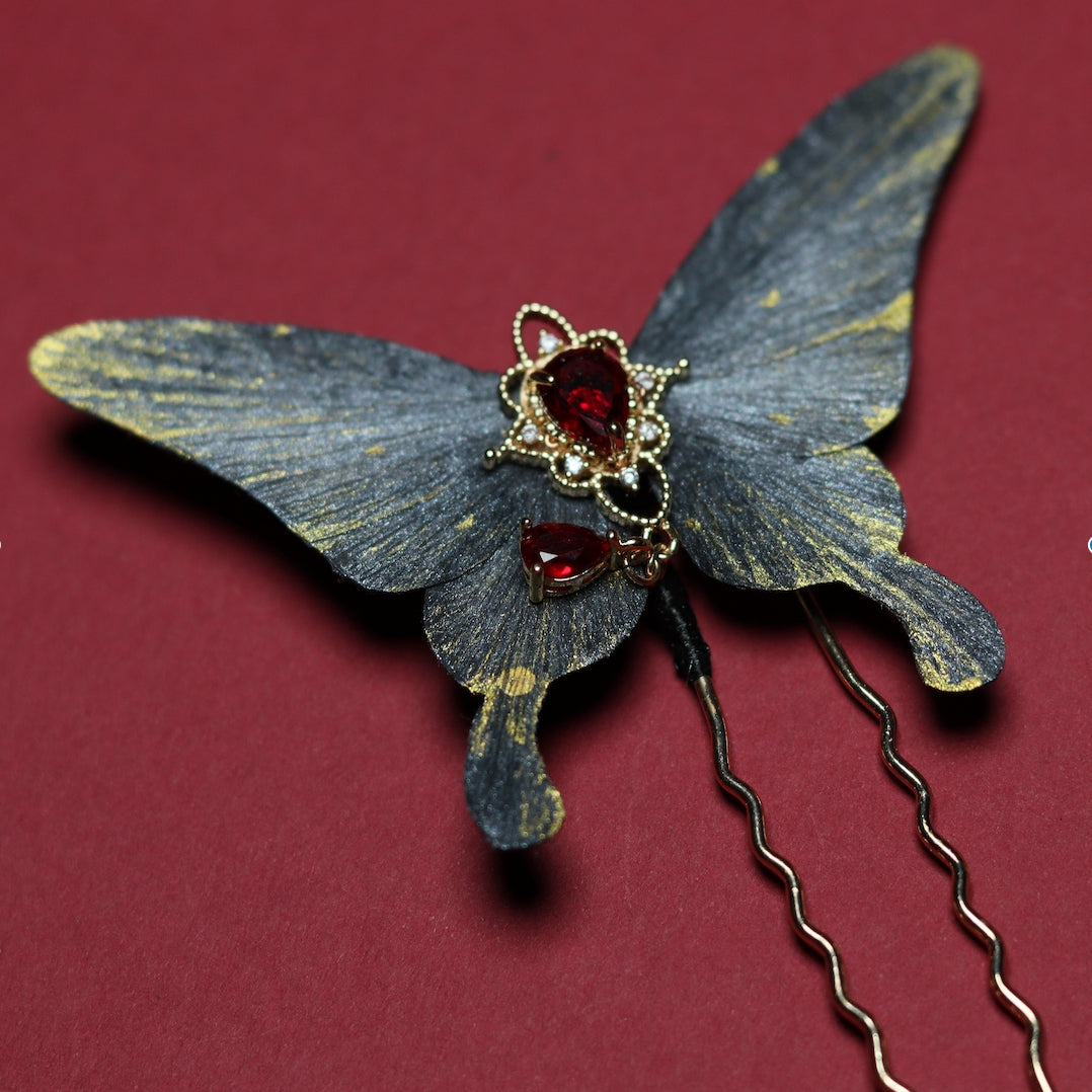 Close-up of a delicate velvet butterfly hairpin featuring a red gemstone centerpiece with golden accents, resting on a crimson background.